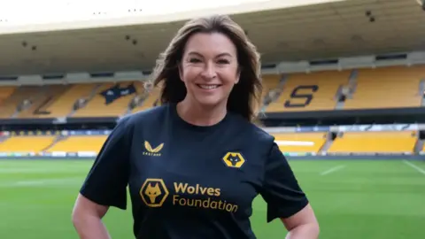 Suzy is on the pitch at Wolves wearing a black Wolves Foundation t-shirt. She is smiling and has long dark hair.