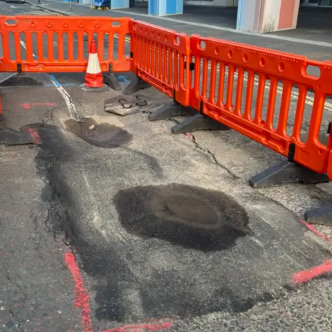 Scottish Water A sinkhole on a tarmac road with two small puddles of water and orange barriers around it