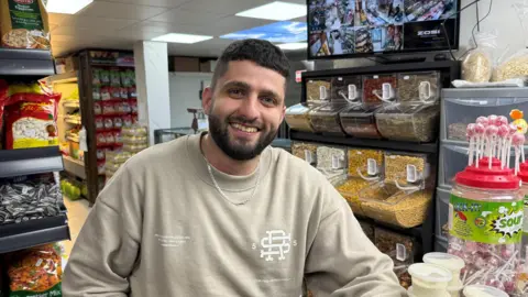 A bearded man in a sweatshirt and chain, smiling at the camera. Behind him are packets of sweets and nuts and pulses and there is a stack of lollipops next to him on the counter he's resting his elbow on.