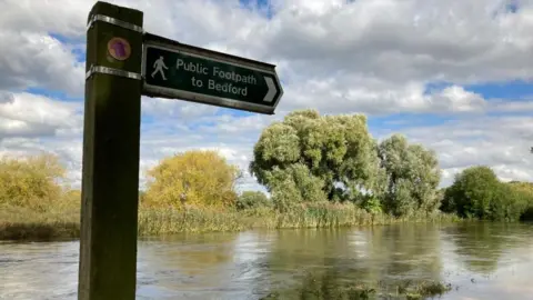 Alex Pope/BBC High water levels on the River Great Ouse at Kempton Mill, Bedfordshire, showing a sign that says Public Footpath to Bedford and trees and bushes along the riverbank.