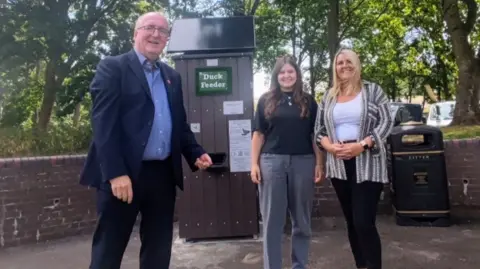 Councillor Richard Morris has balding white hair and glasses and is wearing a navy blue suit with a light blue shirt. He is stood with Charlie Willis and Kirstie Yarwood - Wychavon Parks Team Officers - one has long brown hair and is wearing a black top and grey trousers, while the other has blonde hair and is wearing a white top with a striped shirt open over and black trousers. They are stood with the duck vending machine which has a solar panel on the roof with a wooden panelled frame. A green sign reads duck feeder. 