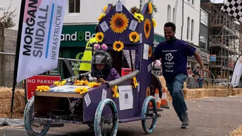 Barrow BID A man in a purple t-shirt pushed a purple painted kart decorated with yellow sunflowers through the street in Barrow. 