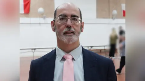 Anthony Felton is looking towards the camera in a school gymnasium w9ht children blurred in the backdground. He is bald on top with close cropped dark hair on the sides. He is wearing glassed and a has a groomed white beard.