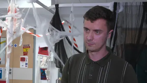 Clinical Skills tutor, Andy Garcia. He has a dark brown, short sleeved polo top, with white and khaki think stripes. He has short dark brown hair. He is inside an escape room. Bandages are hung across the ceiling and there are medical warning posters on the wall.