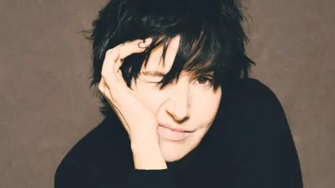 Sharleen with her trademark black hair over her eyes and black clothing. She is smiling as she holds a hand to her face.