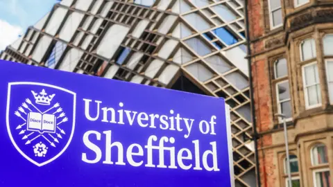 A blue University of Sheffield sign, with the writing in a white font and a university crest to the left. The crest features a book with the writing 'Disce Doce', and is surrounded by arrows, a crown and a white rose. University buildings can be seen behind the sign. 