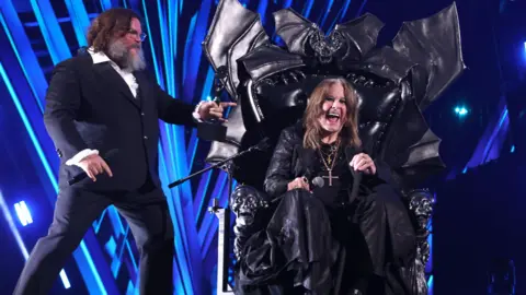 Getty Jack Black is standing on the left, wearing a dark suit with a white shirt. He has shoulder-length brown hair and a beard. Ozzy Osbourne, is sitting in a large black leather bat-shaped chair. He is smiling. He had shoulder-length brown hair long dark clothes and a crucifix around his neck. They are in front of a blue and black backdrop.