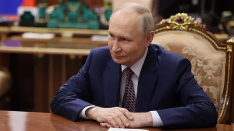 Vladimir Putin sitting at a wooden table, looking off camera, smiling slightly and sitting on a gold chair.