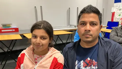 JOANNA TAYLOR/BBC Ten-year-old Keisha is smiling at the camera and sitting next to her Dad, Abhishek, who is looking at the camera with a neutral expression. Keisha is wearing a top patterned with strawberries and a pink and red coat. Her dad is wearing a navy T-shirt and jacket. There are school tables stacked behind them. 