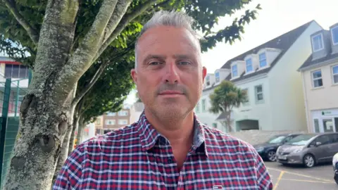 BBC Carl Walker is wearing a red chequered shirt, he has grey hair and is stood under a tree