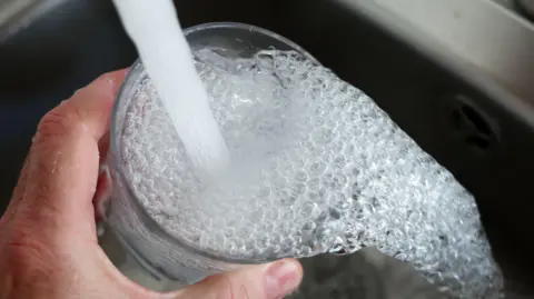 A close-up of a hand holding a glass under a running tap. The water is full of bubbles and is overflowing into a chrome sink.