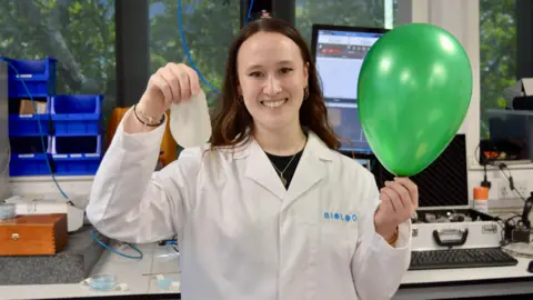 A woman in a lab coat who is holding a green balloon looks at the camera