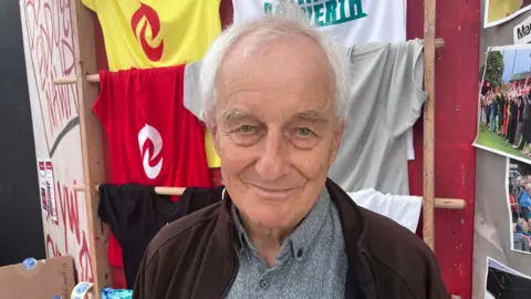Ffred Ffransis pictured in a stall with t-shirts in red and yellow and white displayed on the wall behind him with the Cymdeithas yr Iaith logo shaped like a dragon's tongue. He wears a grey shirt and brown jacket and has grey hair