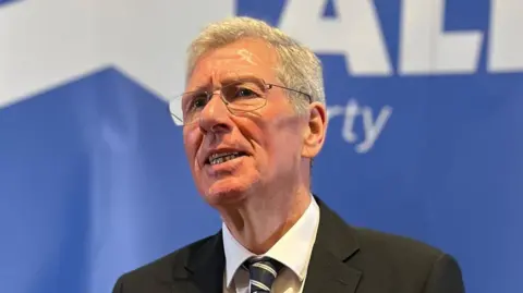 Kenny MacAskill wears a dark suit and striped tie, glasses and has short white hair