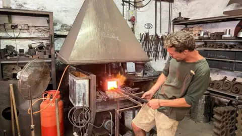 A man, wearing a green t-shirt and beige shorts, holding metal poles which are placed inside a burning furnace. He is in a workshop, surrounded by lots of metal items on shelves. A red gas cylinder is visible to the left of the image, as is an electrical fan.