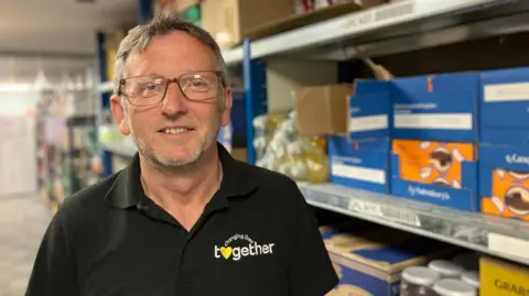 A man in glasses, with short hair and stubble is wearing a black polo shirt with a changing lives together on it. There are racks of food behind him