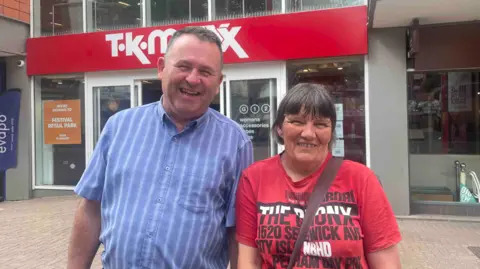 A man with short, mainly dark, hair in a mainly blue shirt with vertical stripes is on the left, next to a woman with dark hair in a red T-shirt with writing on the right. A TK Maxx site is behind them.