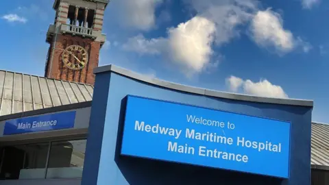 Medway Maritime Hospital Una entrada del edificio con una torre de reloj. Se lee un letrero azul y blanco "Bienvenido a la entrada principal de Medway Maritime Hospital"