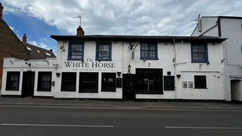 A large, white tow-storey pub beside a two-lane road. Ther are cloudy skies above it. The pub has 12 large windows and two doors. The pub's name, "The White Horse" is written on the front.