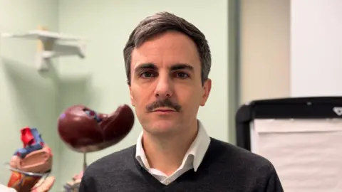 Tom Dolphin, a man with brown hair and a moustache, wears a grey jumper and a white shirt.