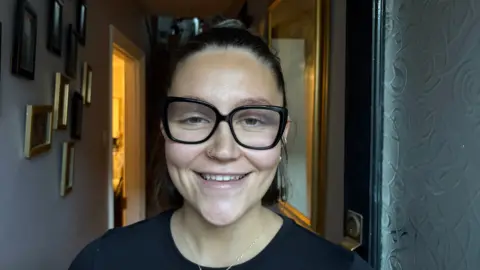 Martin Heath/BBC Grace Connolly with dark hair tied back and wearing black-framed glasses. She is smiling at the camera and wearing a black T-shirt. She is standing in the hallway of a house, with pictures in small frames to her right and a larger frame to her left.
