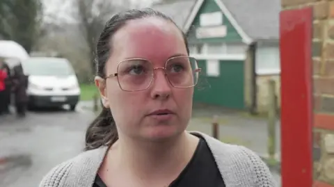 BBC worn a female glasses, a black top and a gray cardigan. There is a white van in the background. 