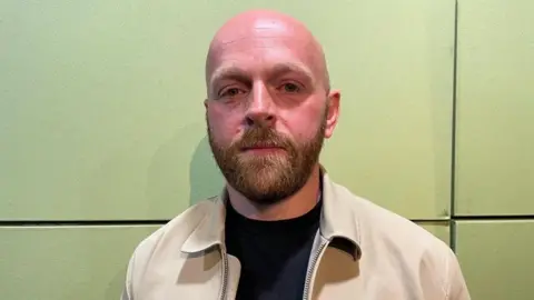 Sean Egan, pictured with a neutral expression on his face. He is bald but has a dark blonde beard, he wears a black t-shirt underneath a beige jacket.



