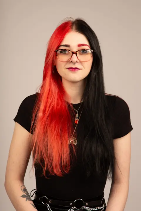 Poppy Bellamy Poppy Bellamy - a woman with black and red coloured hair and eyebrows. She wears black and red framed glasses and a black T-shirt.