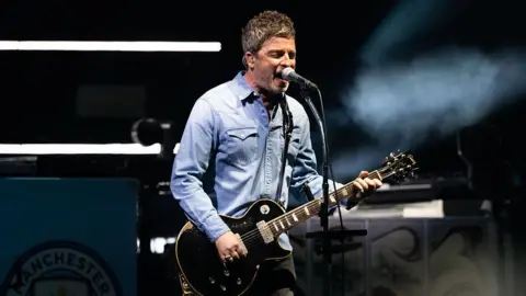 Noel Gallagher on stage in Cardiff singing and playing black guitar. He wears a denim shirt and black jeans