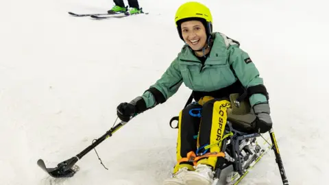 Ellie skiing wearing a yellow helmet, green ski jacket, yellow and black trousers and white trainers. She holds two ski poles out to either side and has a big smile