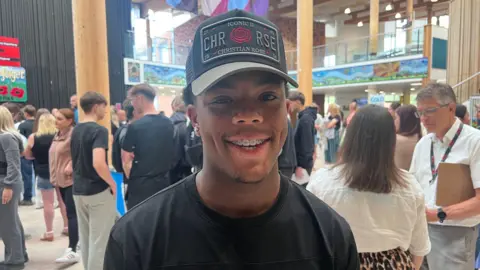 A 16-year-old boy is smiling wearing a black cap and braces and a black t-shirt. There are crowds of people behind him.