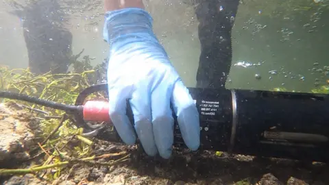 A hand wearing a blue glove holds a long black cylinder underwater with a cable attached to the rear 