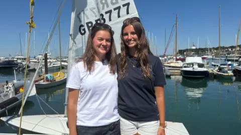 Image shows Freya and Maddie smiling at the camera. Freya is wearing a white t-shirt and black denim shorts. She has brown hair and sunglasses on her head. She is stood with her arm around Maddie, who has long brown hair. She also has sunglasses on her head. She has a blue t-shirt on and white denim shorts. Behind them, is their boat called 'Fiasco'. You can see part of Cowes harbour behind them, with blue skies and blue water.