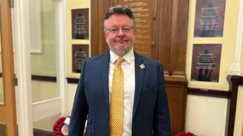 BBC Man in blue suit jacket white shirt yellow tie grey beard and glasses stands in a hallway in front of wooden war memorial plaque 