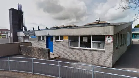 A school building with a road and concrete area in front. The door and window frames are painted blue and there are pictures and notices stuck to the inside of windows