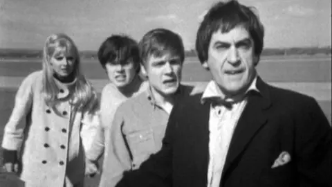 Black and white screen grab image of four people looking startled on a runway