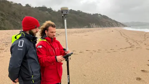Two men stand on the sandy beach in Slapton Devon after Storm Ingrid. They are measuring how the height of the beach has changed using digital equipment. There are footprints in the sand into the distance and a few silated houses at the top of the shrub covered cliffs behind the beach. 