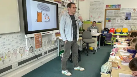 Author and illustrator Tom McLaughlin stands in front of a smart board with a sketch of a dog and an orange cat on it. The children sit at desks in front of him, learning to draw their own animals, for their own comic books that they are making.