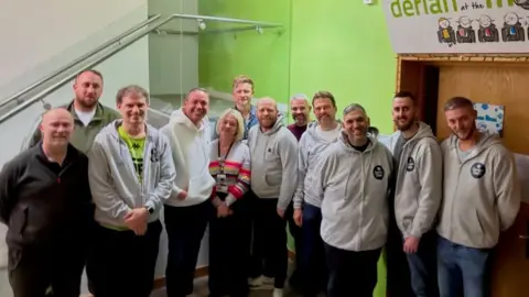 Derian House handout A group of bereaved dads - some wearing grey hoodies with a Derian Dads logo on it - and staff outside an in-house cinema inside Derian House Children's Hospice for the premiere of a documentary. They are all smiling.