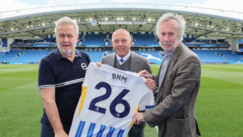 Three men stood in a football stadium. They are holding a white and blue football shirt, with "BHM" and "26" on the back.