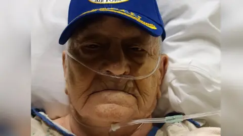 Handout An elderly man in a hospital bid with an American air force cap on in blue and gold and a number of tubes attached to him