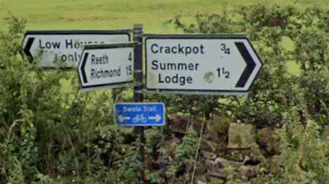 The image shows a rural road junction with several directional signs set against green fields. The signs point towards places including Crackpot, Summer Lodge, Low Houses and Reeth. In the background are stone walls, scattered sheep grazing, and a couple of small stone buildings.