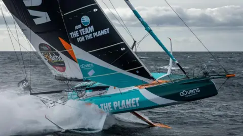 Pierre Bouras A racing yacht with a white and blue hull and black and white sails going fast with spray behind.