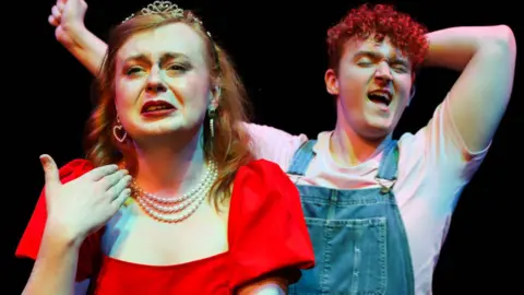 Amanda Doherty Rose and Conor, with a pitch-black background behind them. Rose is wearing a red dress, while Conor is wearing a white t-shirt and blue overalls.