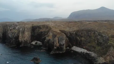 The site is an area of steep sea cliffs. It is a rugged, bare landscape with hills in the background.