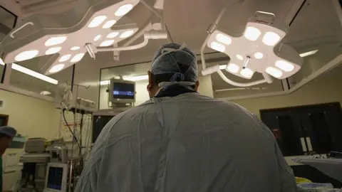 Getty Images Surgeon seen from the back in operating theatre under bright overhead lights in a surgical gown