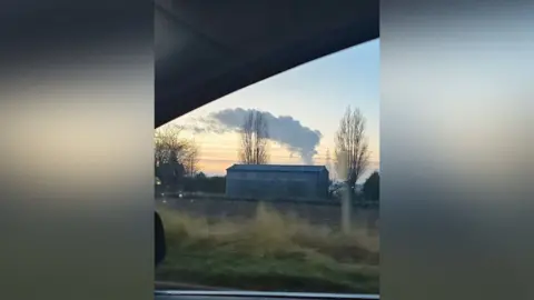 Debbie Peters The photo has been taken outside of a car window. It shows a visible cloud plume that is coming from behind a large farm-style warehouse. This is situated behind a field that crops grow on. There are various trees in the distance too. 