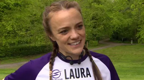 Laura Aiken, a woman with long, fair hair tied back in two plaits, speaks to a BBC TV crew at the Stormont estate. She is wearing a two-tone sports top with a white chest and purple sleeves, and a set of black headphones. 