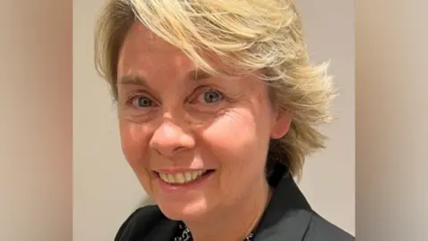 Sam Ridding, a woman with blonde hair and wearing a black shirt is smiling. She is standing against a light-coloured background.