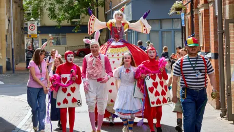 Performers dressed up as Alice in Wonderland characters, including Alice, The White Rabbit and the White Queen walking on the streets of Oxford. It is sunny.